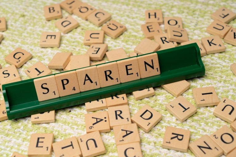 Auf einer gehäkelten hellgrünen Decke ist das Wort "Sparen" mit Scrabble-Buchstaben gelegt und rundherum liegen weitere Buchstaben.